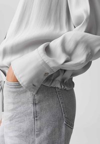 Hellgraue Bluse mit langen Ärmeln, aus einem weichen, glatten Material und mit gerafftem Bund; sie hat eine Knopfmanschette und ein minimalistisches Design, kombiniert mit hellgrauen Jeans.