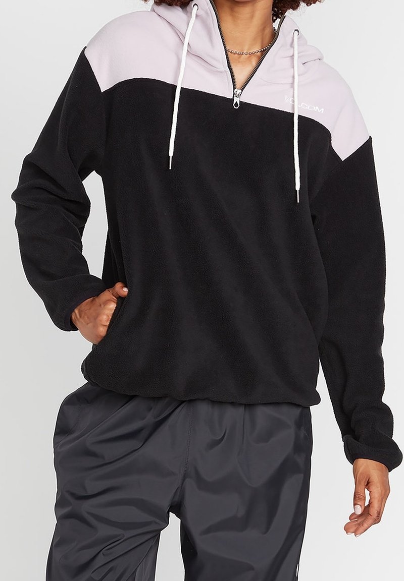 Personne portant un pull polaire noir avec un empiècement rose clair, des cordons blancs et un pantalon gris foncé sur fond uni.