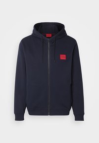 Sudadera con cremallera de color azul marino, con capucha ajustable y parche rectangular rojo con la etiqueta "HUGO" en el lado izquierdo del pecho.
