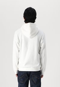 Sweatshirt à capuche blanche avec une coupe décontractée, des poignets et une taille côtelés, porté par-dessus un jean bleu foncé, vu de dos.