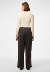 Kobieta z krótkimi brązowymi włosami, zwrócona tyłem, ubrana w kremowy prążkowany top z długim rękawem i ciemnobrązowe szerokie spodnie.