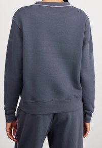 En person som bär en mörkgrå långärmad sweatshirt och matchande träningsbyxor, sedd bakifrån mot en neutral bakgrund.