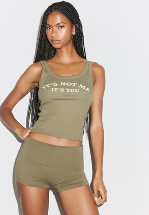 Jeune femme aux longs cheveux tressés porte un débardeur vert olive avec le texte « "IT'S NOT ME IT'S YOU" » et un short assorti sur un fond clair uni.