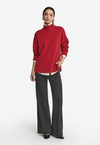 Roter Strickrollkragenpullover mit gerippten Bündchen und Saum, kombiniert mit weitgeschnittenen, dunkelgrauen Hosen und schwarzen spitzen Schuhen.