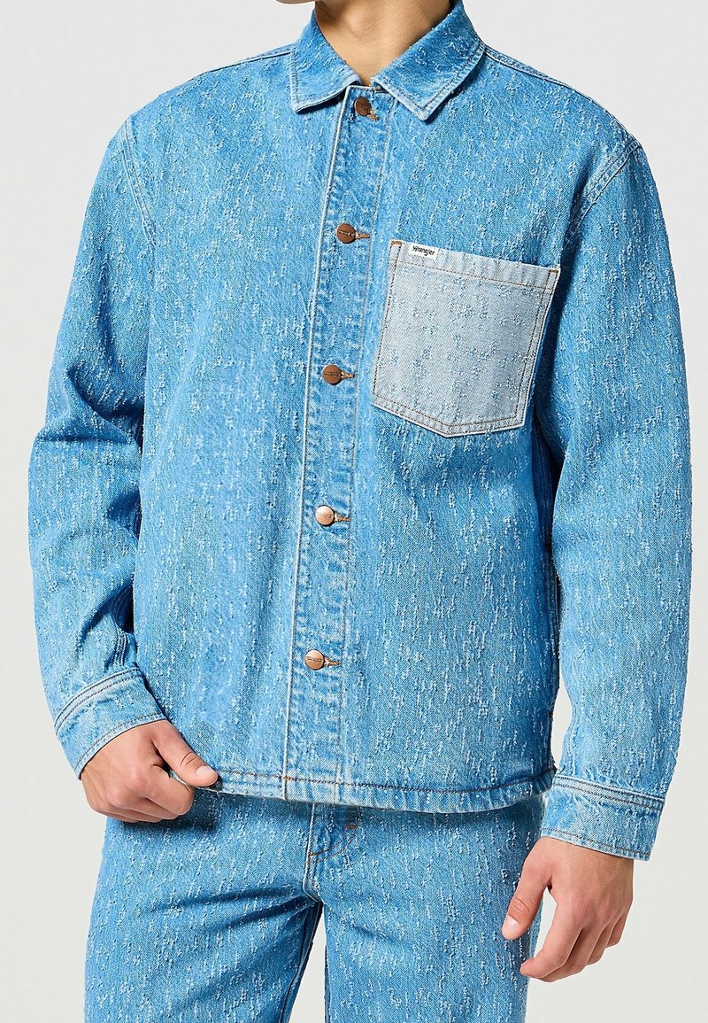 Giacca di jeans azzurro chiaro con finitura texturizzata, caratterizzata da un colletto a punta, bottoni frontali e una tasca applicata sul lato sinistro del petto.