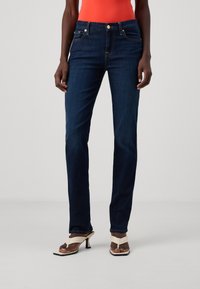 Σκούρα μπλε τζιν παντελόνια με εφαρμοστό μέση, ίσια γραμμή, χρυσό κουμπί και αντίθετη ραφή. Φορεμένα με λευκά ψηλοτάκουνα σανδάλια.