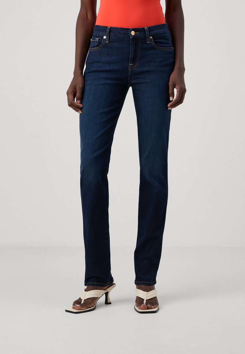 Σκούρα μπλε τζιν παντελόνια με εφαρμοστό μέση, ίσια γραμμή, χρυσό κουμπί και αντίθετη ραφή. Φορεμένα με λευκά ψηλοτάκουνα σανδάλια.