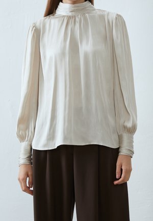 Femme portant une blouse en soie blanche à manches longues avec un col haut et une coupe ample, associée à un pantalon large marron foncé.