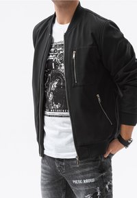 Black bomber jacket with zip pockets, ribbed cuffs, and a smooth texture, layered over a graphic T-shirt and distressed gray jeans.