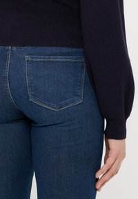 Δερμάτινο τζιν σε σκούρο μπλε χρώμα με λεία υφή. Διαθέτει δύο πίσω τσέπες με λευκή ραφή. Συνδυάζεται με μια σκούρα πλεχτή μπλούζα.