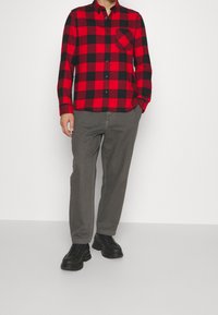 Chemise en flanelle à carreaux rouges et noirs avec fermeture à boutons et une poche poitrine, associée à un pantalon gris et des chaussures noires à lacets.