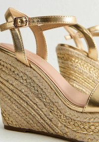Sandali a cuneo dorati con cinturini metallici, dettagli in juta intrecciata e superficie texturizzata; il sottopiede rosa pallido contrasta con il design.