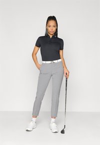 Μαύρο polo μπλούζα με γιακά και κουμπιά, συνδυασμένο με ανοιχτό γκρι tailored παντελόνι. Μια λευκή ζώνη και παπούτσια ολοκληρώνουν την εμφάνιση. Κράτησε ένα γκολφ κλαμπ στο ένα χέρι.