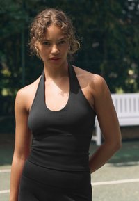 Top nero a canotta con scollo a U e texture a coste, design aderente e tessuto liscio, indossato con un capo inferiore abbinato.