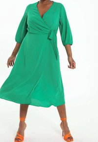 Grünes Wickelkleid mit Dreiviertel-Puffärmeln, Bindegürtel in der Taille und fließendem Rock. Getragen mit orangefarbenen Riemchensandalen.