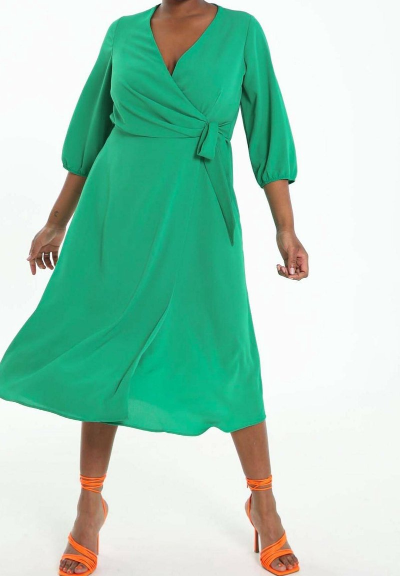 Grünes Wickelkleid mit Dreiviertel-Puffärmeln, Bindegürtel in der Taille und fließendem Rock. Getragen mit orangefarbenen Riemchensandalen.