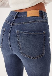 Džíny s vysokým pasem v modré denimové barvě s oranžovým prošitím, dvěma zadními kapsami a koženou nášivkou se značkou na pase.