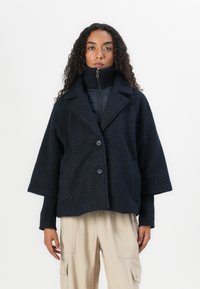 Navy blue textured wool blend jacket with a collar, two buttons, and a boxy fit. Worn over a zipped high-neck inner layer.