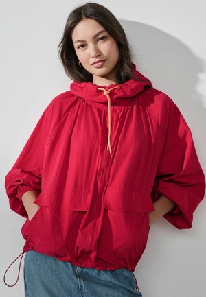 Junge Frau mit dunklen Haaren trägt eine leuchtend rote Kapuzenjacke mit Kordelzügen und blaue Jeans, stehend vor einem schlichten hellen Hintergrund.