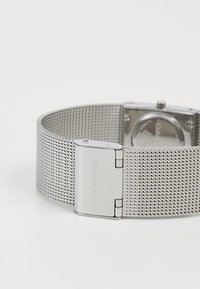 Srebrna metalna mrežasta narukvica za sat s graviranim kopčom Calvin Klein, djelomično vidljiv stražnji dio sata na bijeloj površini.