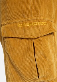 Buks i corduroy i gyldengul, med lodrette ribber, en frontlomme med klap og broderet brandlogo i matchende farve.