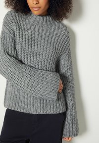 Femme portant un épais pull en maille côtelée gris avec des manches longues et un pantalon noir, debout devant un mur blanc uni.