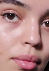 Primer plano de un rostro con piel suave y clara, labios carnosos con un acabado brillante y ojos marrón oscuro enmarcados por pestañas y cejas naturales.