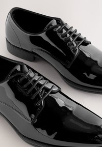 Paire de chaussures habillées en cuir verni noir poli avec lacets fermés sur fond clair.