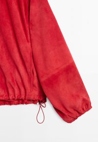 Rukáv červené semišové bundy s nařaseným elastickým lemem a nastavitelným stahovacím šňůrkem na spodním okraji na bílém pozadí.