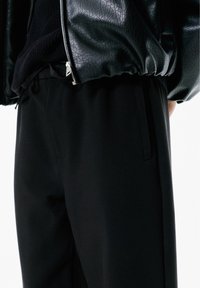 Pantalon noir en tissu lisse, avec des poches latérales, une coupe décontractée et une légère brillance. Associé à une veste zippée noire texturée.