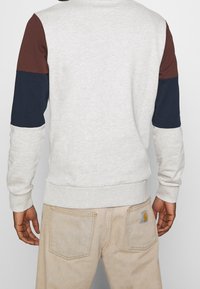Grå sweatshirt med burgunder och marinblå ärmar, texturerad tyg, ribbad mudd och nederkant, i kombination med beige jeans med bakfickor och logotyp.