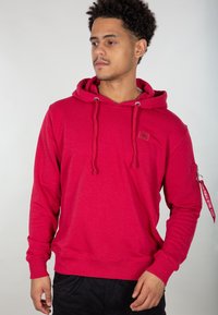 Felpa con cappuccio rosso realizzata in tessuto morbido, con cordini, polsini a costine e tasca con zip dotata di una fascia decorativa.