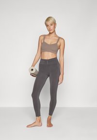 Sujetador deportivo marrón claro con tirantes ajustables y leggings de talle alto grises. Las piernas tienen costuras acentuadas, y se sostiene una esterilla de yoga enrollada en una mano.