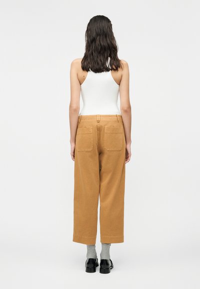 Γυναίκα με σκούρα μαλλιά μήκους μέχρι τους ώμους, που φοράει λευκό τοπ χωρίς μανίκια, παντελόνι σε ανοιχτό καφέ με φαρδιά μπατζάκια, γκρι κάλτσες και μαύρα παπούτσια, στέκεται με την πλάτη γυρισμένη.