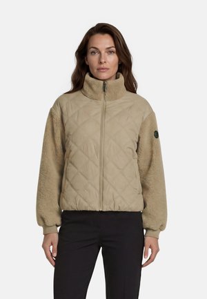 Vrouw draagt een beige doorgestikte jas met fleece mouwen en kraag, staand tegen een effen witte achtergrond.