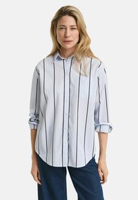 Light blue button-up shirt with vertical black and blue stripes, featuring a chest pocket and rolled-up sleeves. Worn with dark jeans.