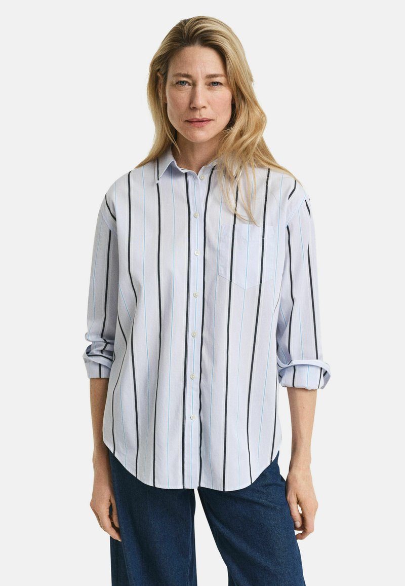 Light blue button-up shirt with vertical black and blue stripes, featuring a chest pocket and rolled-up sleeves. Worn with dark jeans.