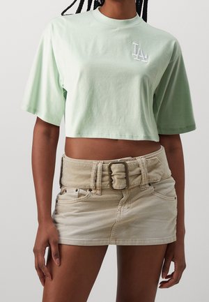 T-shirt verde menta corta con logo LA, abbinata a una minigonna in velluto a coste beige con cintura larga e tasche frontali.