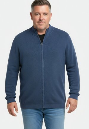 Hombre con un suéter azul texturizado con cremallera y vaqueros azul claro, de pie frente a un fondo blanco liso.