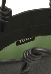 Borsa nera a trama con interno verde, dotata di chiusura a cerniera e dettagli con manici annodati. Etichetta interna visibile.