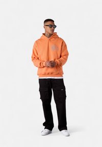 Sweat-shirt à capuche orange avec poche frontale, design brodé, associé à un pantalon cargo noir et des baskets blanches. Lunettes de soleil portées.