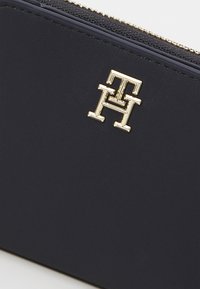 Portefeuille en cuir marine avec un accent logo en ton or, fermeture éclair et finition texturée sur le bord supérieur.