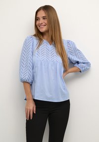 Chemisier en coton bleu clair avec un décolleté en V, des manches bouffantes et des détails en œillet à motif chevron. Caractérisé par une coupe décontractée et une taille froncée.