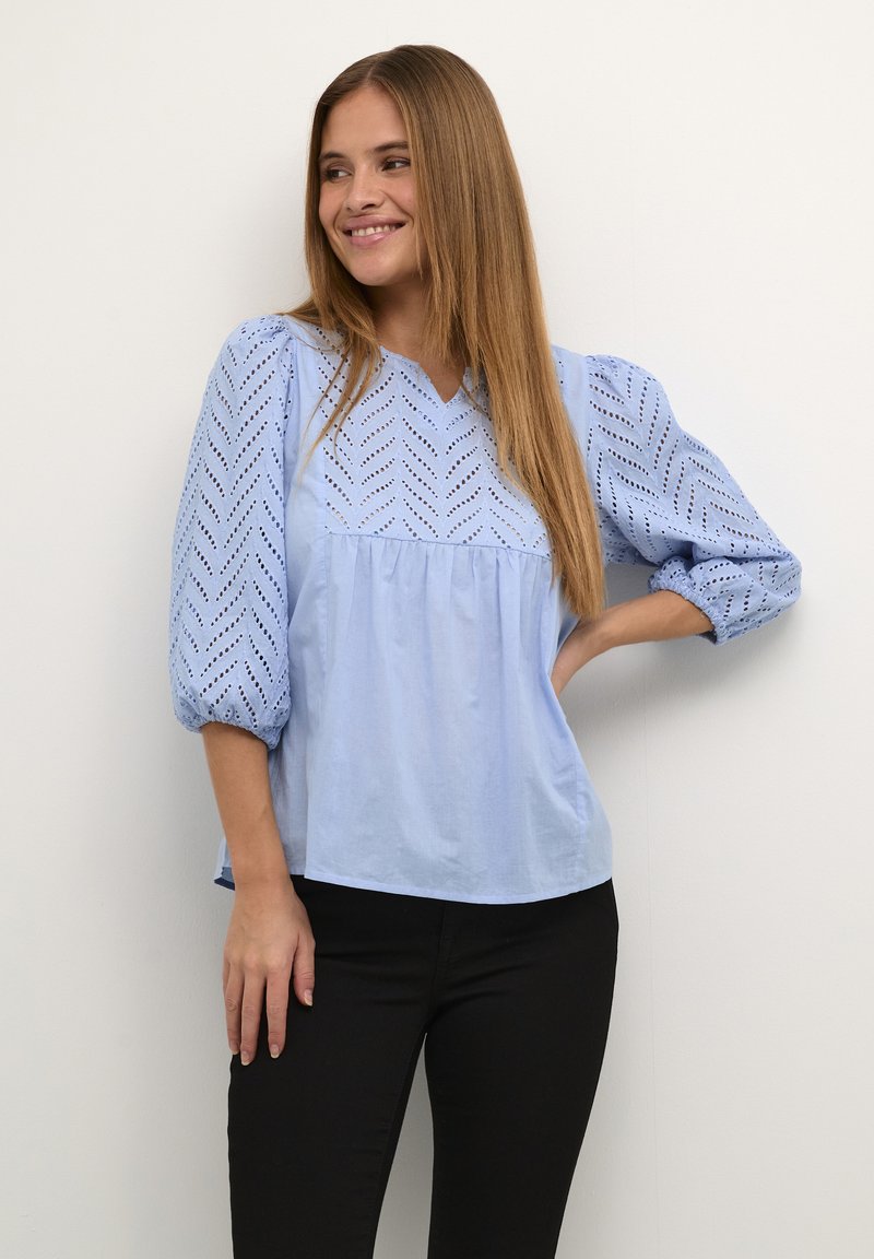 Chemisier en coton bleu clair avec un décolleté en V, des manches bouffantes et des détails en œillet à motif chevron. Caractérisé par une coupe décontractée et une taille froncée.