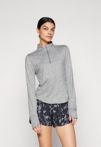 Graues, langärmliges Sportoberteil mit Kragen und halbem Reißverschluss, kombiniert mit dunkel gemusterten Shorts mit einem Batikdesign. Glatte Textur.
