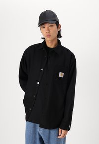 Zwarte button-up shirt gemaakt van stof met een gladde textuur, met een borstzak en metalen knopen, gecombineerd met een zwarte pet.