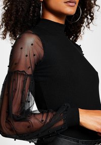 Maglione a collo alto a coste nere con maniche a sbuffo trasparenti decorate con perline nere e polsini arricciati.