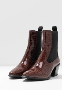 Botas de tobillo de charol marrón con textura de cocodrilo, puntera afilada, paneles laterales elásticos negros y un tacón corto y grueso.