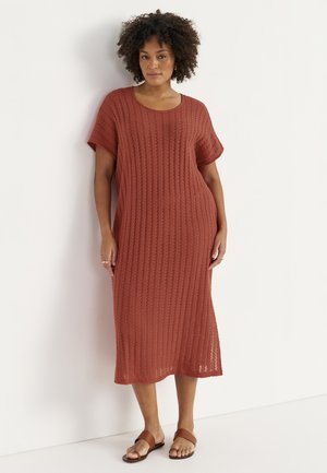 Kvinde med krøllet hår iført en rustfarvet, knælang strikket kjole og brune sandaler, stående mod en hvid væg.