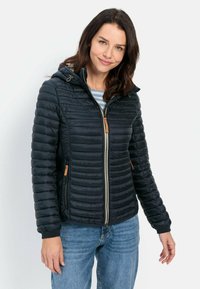 Gewatteerde zwarte jas met een rits aan de voorkant, capuchon en geribbelde manchetten. Bevat tan accenten en een diagonaal quiltpatroon. Draagt over een gestreepte top.